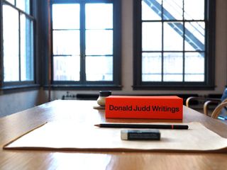 A book with a red cover says 'Donald Judd Writings'. It's photographed on a work desk, next to paper, a pen, and an eraser.