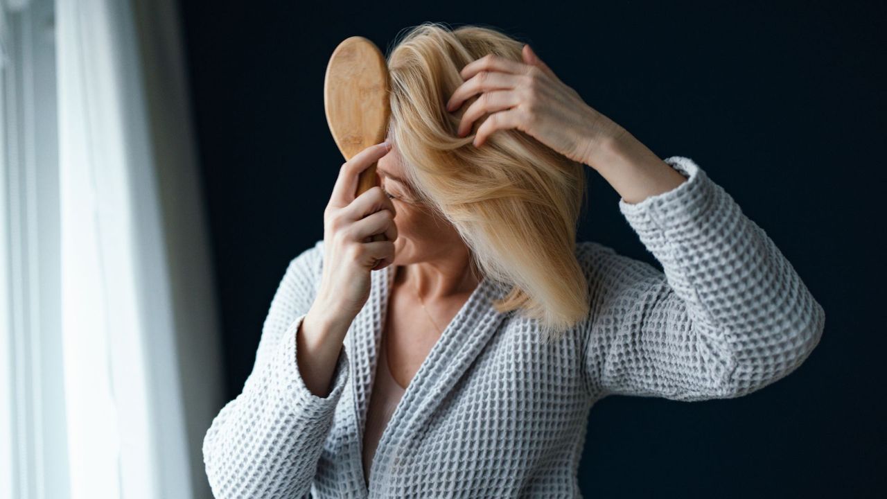 Woman brushing through hair with hairbrush wearing dressing gown, going through hair loss and menopause