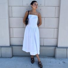 Dominique wears white linen dress with black shoulder bag and black mesh flats while leaning on a wall and posing.