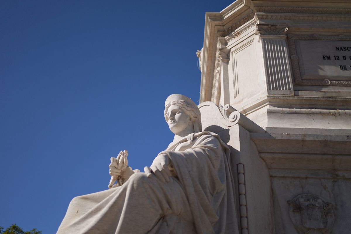 White stone seated statue photographed from below against a deep blue sky and an ornate monument.