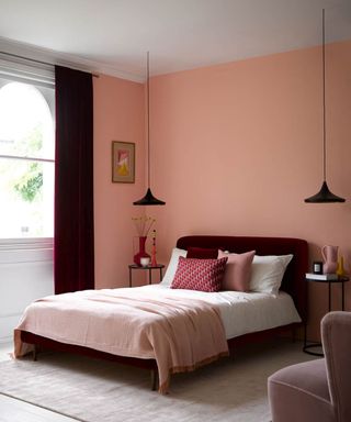 Peach walls with burgundy contrast velvet bed and full length drapes, with tonal bedding, pillows and accessories, and black pendant lights over bedside tables.