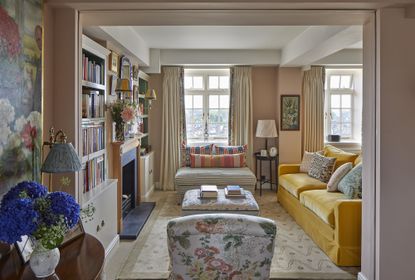 living room space with yellow couch, floral armchairs, coral walls, bookcases, artwork, drapes, patterned cushions, rug