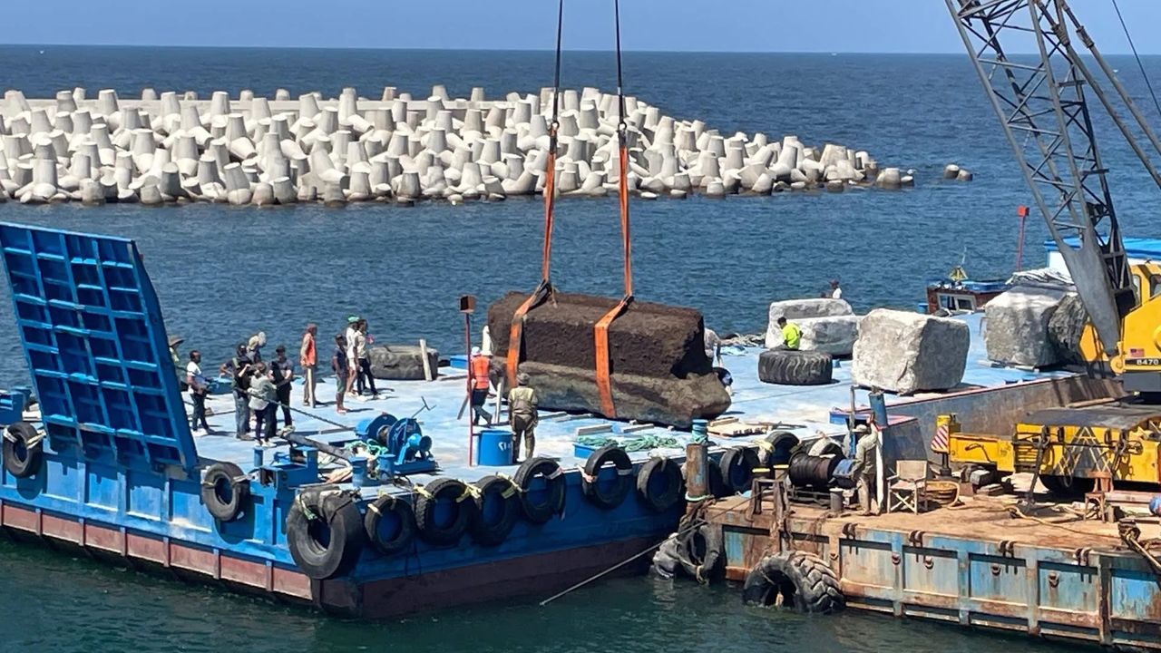 a crane lowers a large stone block onto a platform in the ocean as people stand and watch