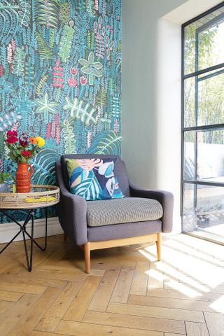 Corner in kitchen with blue-green patterned Lucy Tiffney wallpaper, herringbone wood floor and grey armchair