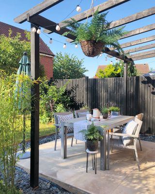 a painted black pergola with a white dining set underneath, sat on a paved beige patio, with a black fence next to it