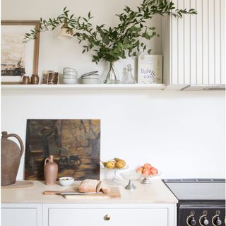 white shelf with plant in a vase, artwork and bowls above a white worktop with a brown vase, painting and chopping board