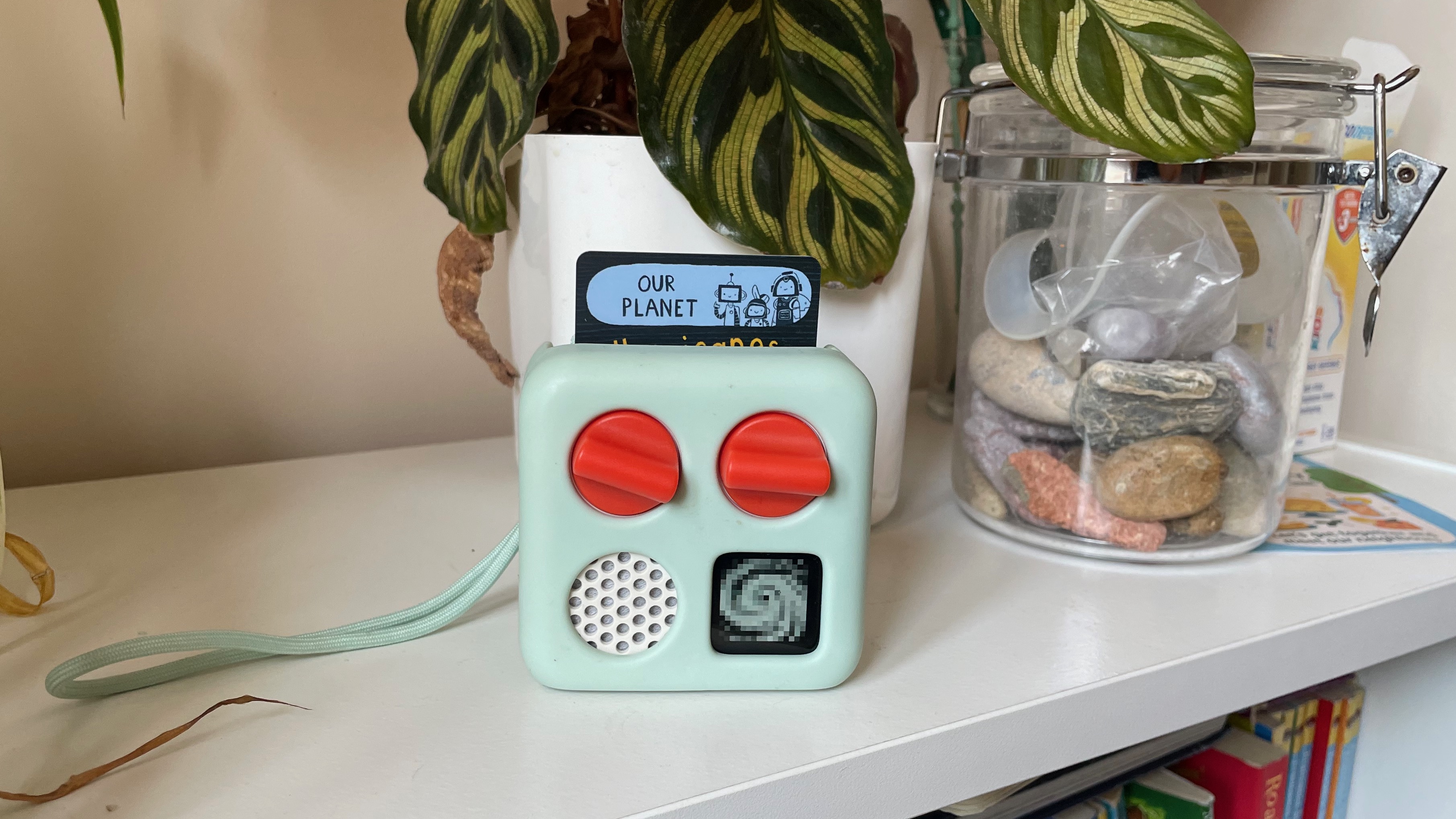 A Yoto Mini player in a green case on top of a bookcase in front of a pot plant and a clear container of stones. The card in the Yoto Mini reads 'Our Planet', and on-screen is an 8-bit graphic of a tornado.