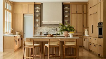 Spacious modern kitchen with light oak cabinetry, a large central island with three cane-back barstools, a marble backsplash, and a prominent white range hood.