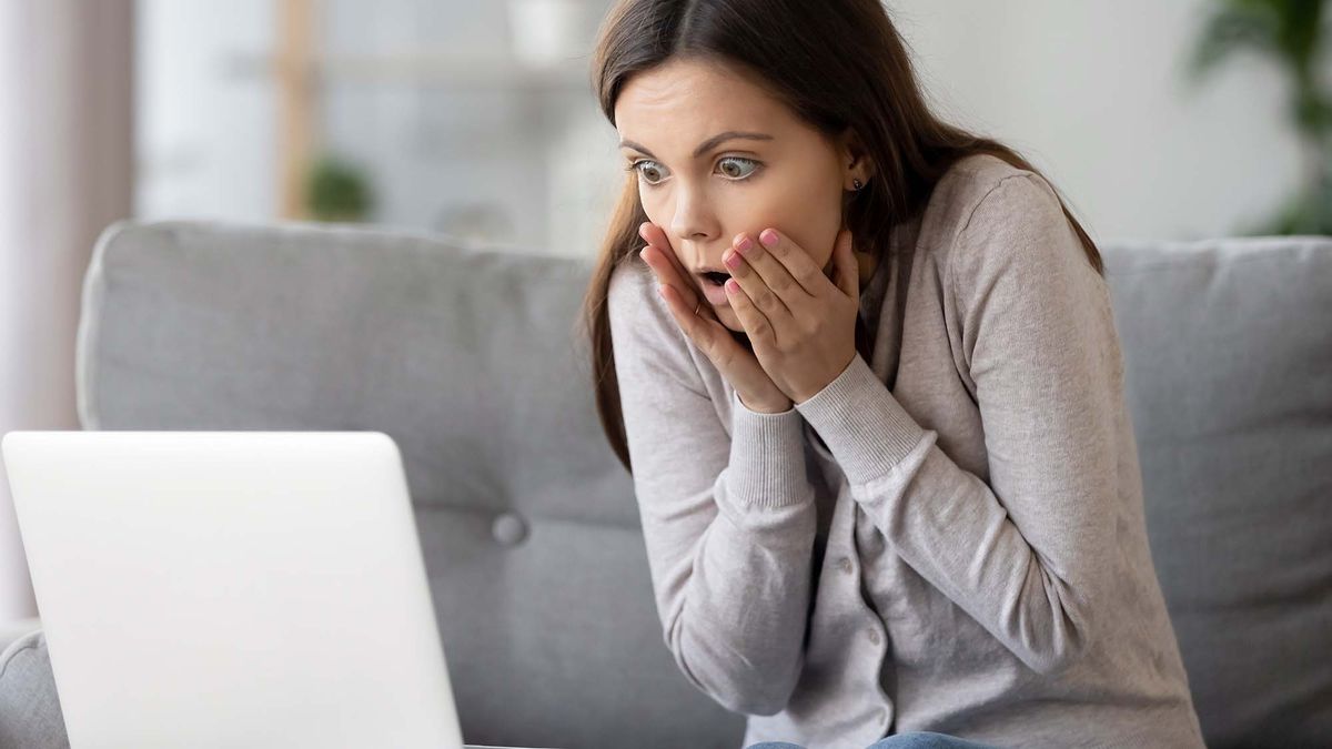 A shocked girl sitting on sofa at home looking on laptop screen