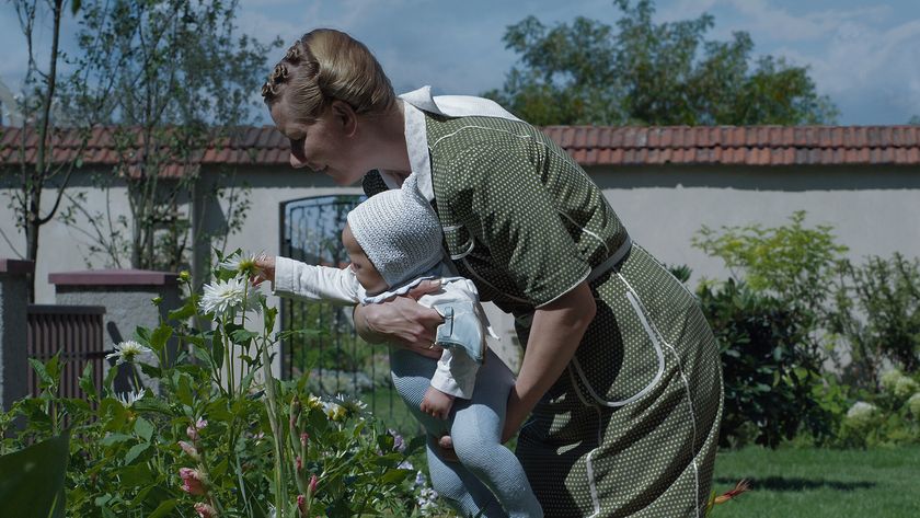 A woman in a garden holding a young baby as it reaches out to touch a flower.