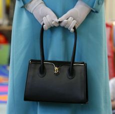 Queen Elizabeth II wearing a blue coat and holding a black purse behind her while wearing white gloves