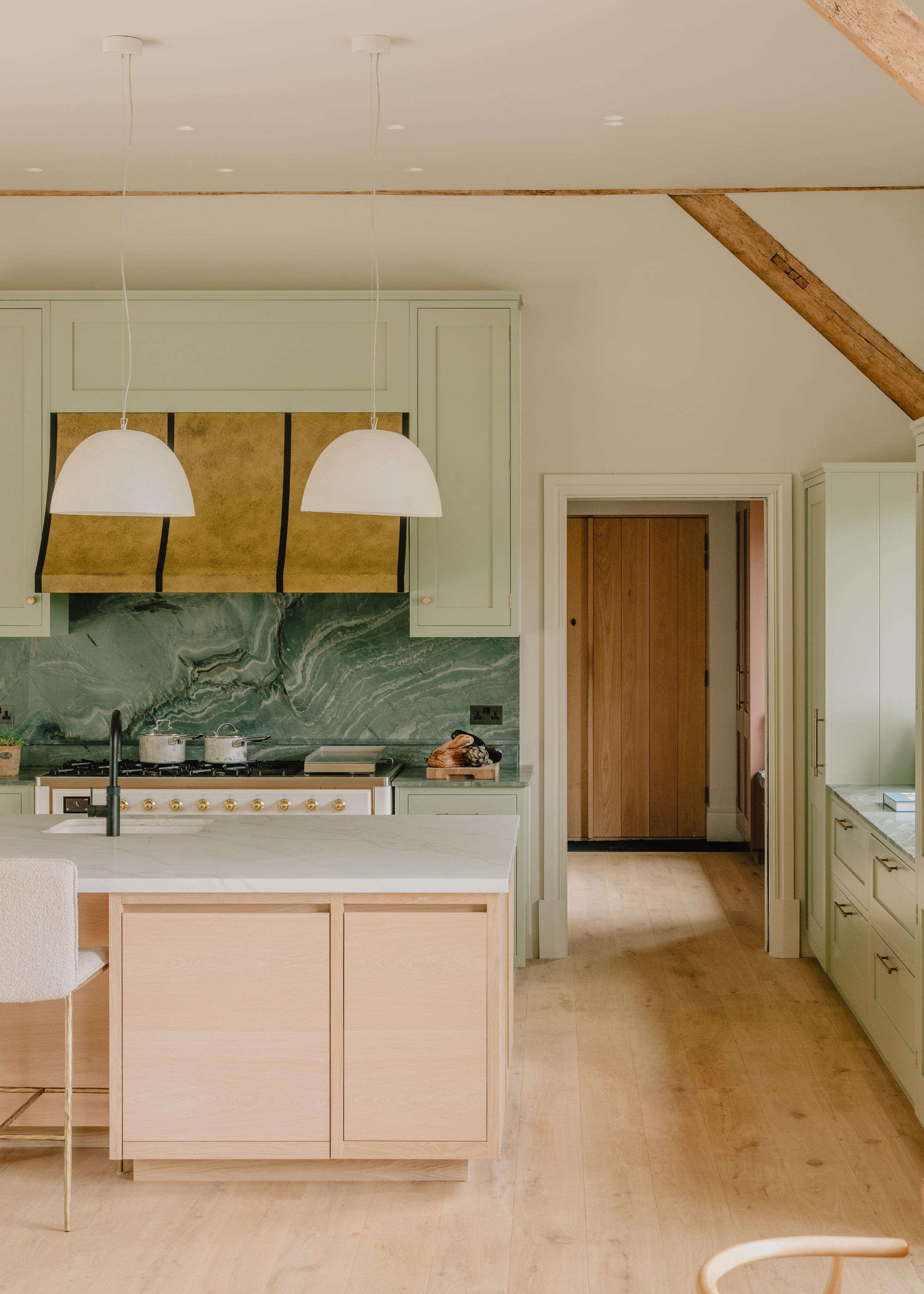 A pastel pink and green modern kitchen with an island and white bar chairs tucked underneath and white pendant light above. The back splash on the w=back wall is in green stone and the extractor hood is brass