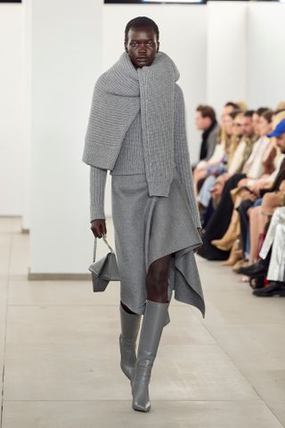 Michael Kors fall/winter 2024 model wears a gray sweater tied over another gray sweater with a skirt and knee-high boots.