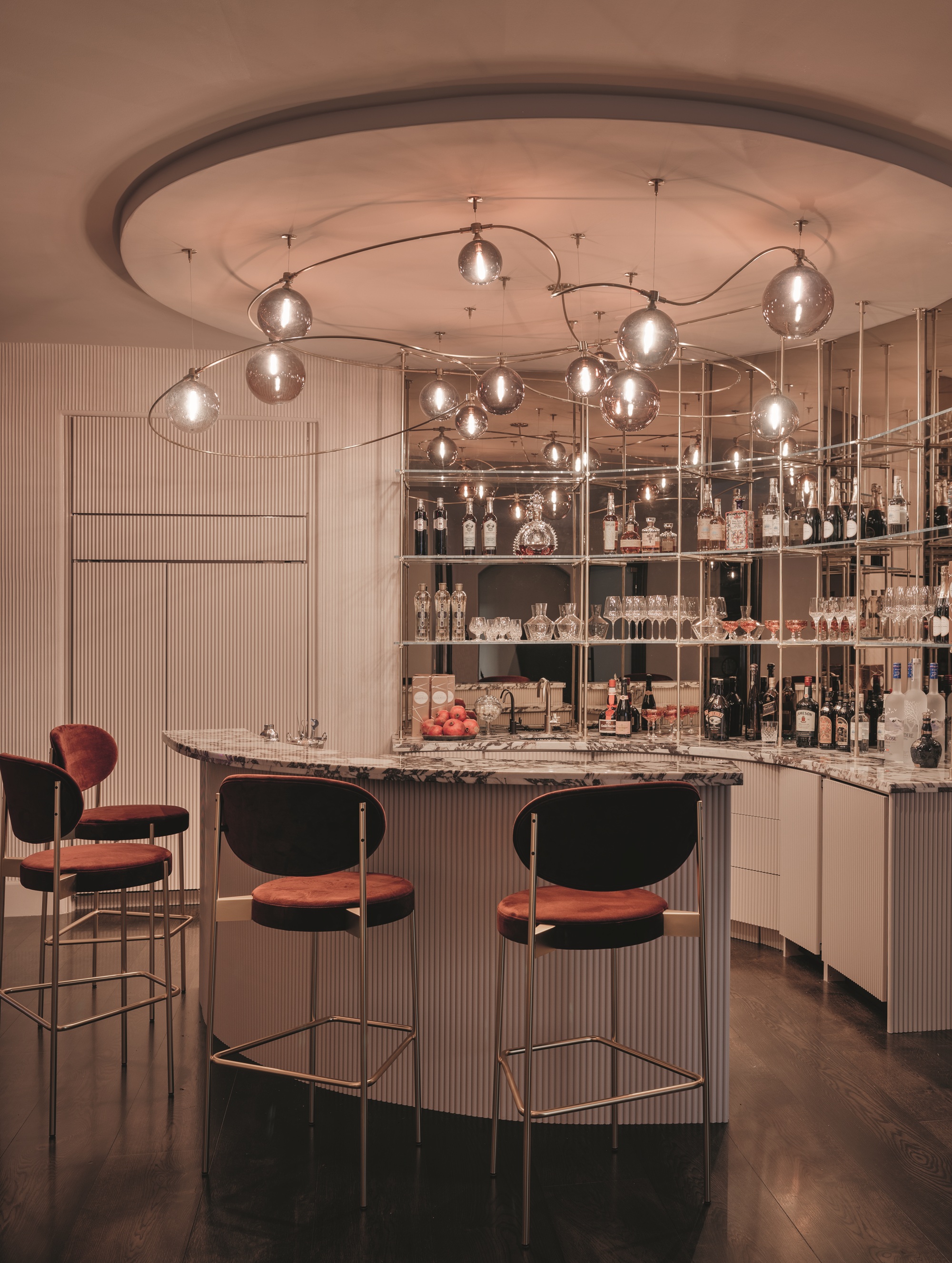 home bar area with counter, fluted detail, mirrored shelves styled with bottles and glassware, custom pendant light, fluted wall paneling, and four pink velvet bar stools