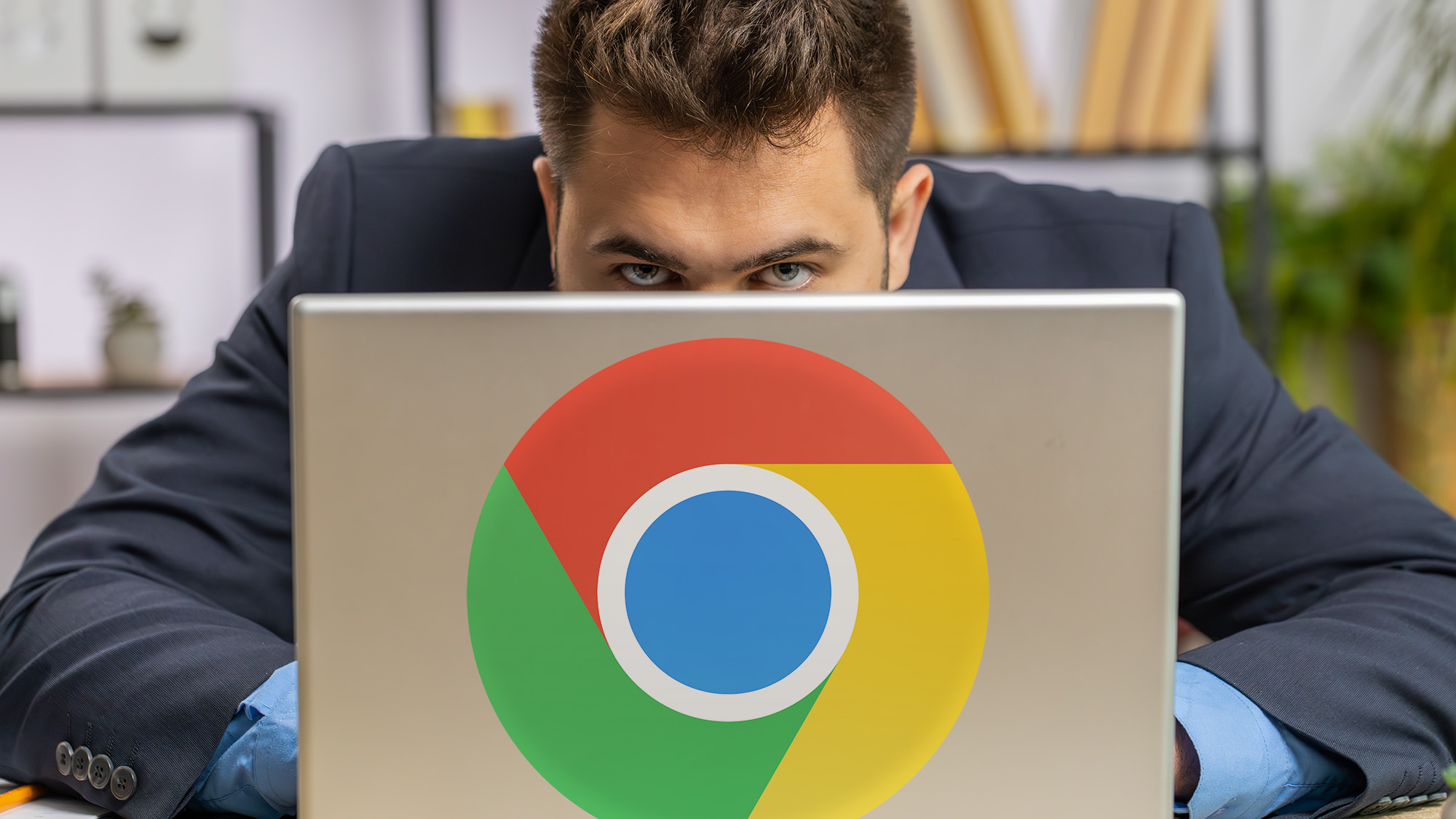 A man in a suit peers over a laptop displaying a large Chrome logo. The setting is an office.