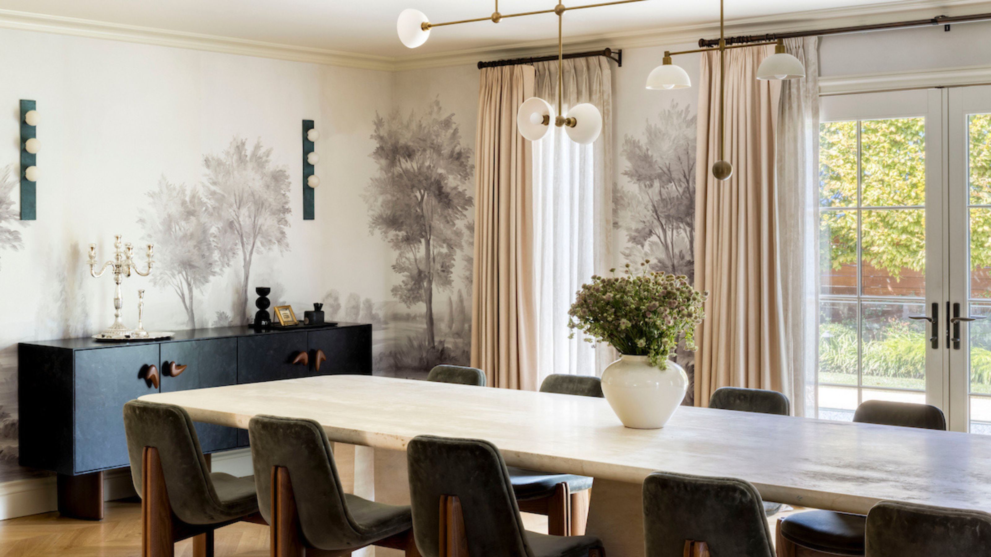 A dining room with a large table in the middle and chairs surrounding it. There is also a hand-painted mural encapsulating the space as well as a side table with storage space inside. You can see a slight view of the outside of the home leading to the garden area.