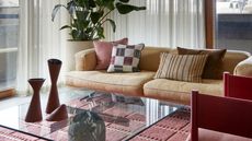 A yellow sofa in a living room with floor-to-ceiling windows, a pink rug, a glass coffee table, and a plant in the corner.