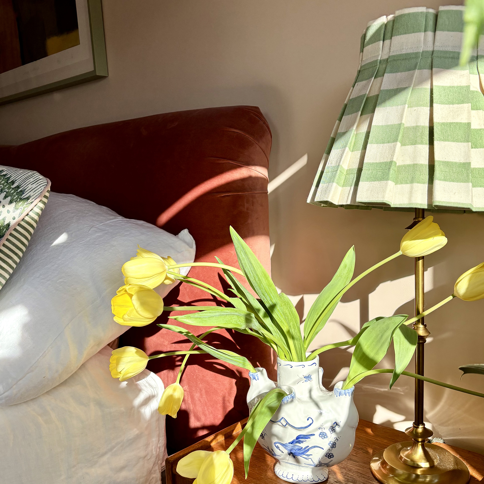 Detail image of a bedside table with a lamp with green striped shade, and a vintage jug filled with yellow tulips. A terracotta velvet headboard is visible behind