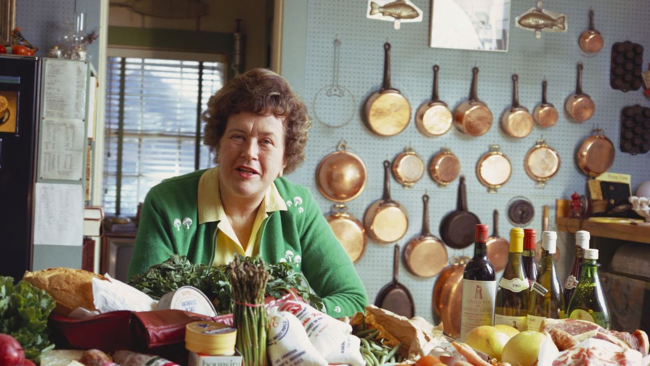 Julia Child in her kitchen with copper pans hung on the wall behind her and fresh produce on table