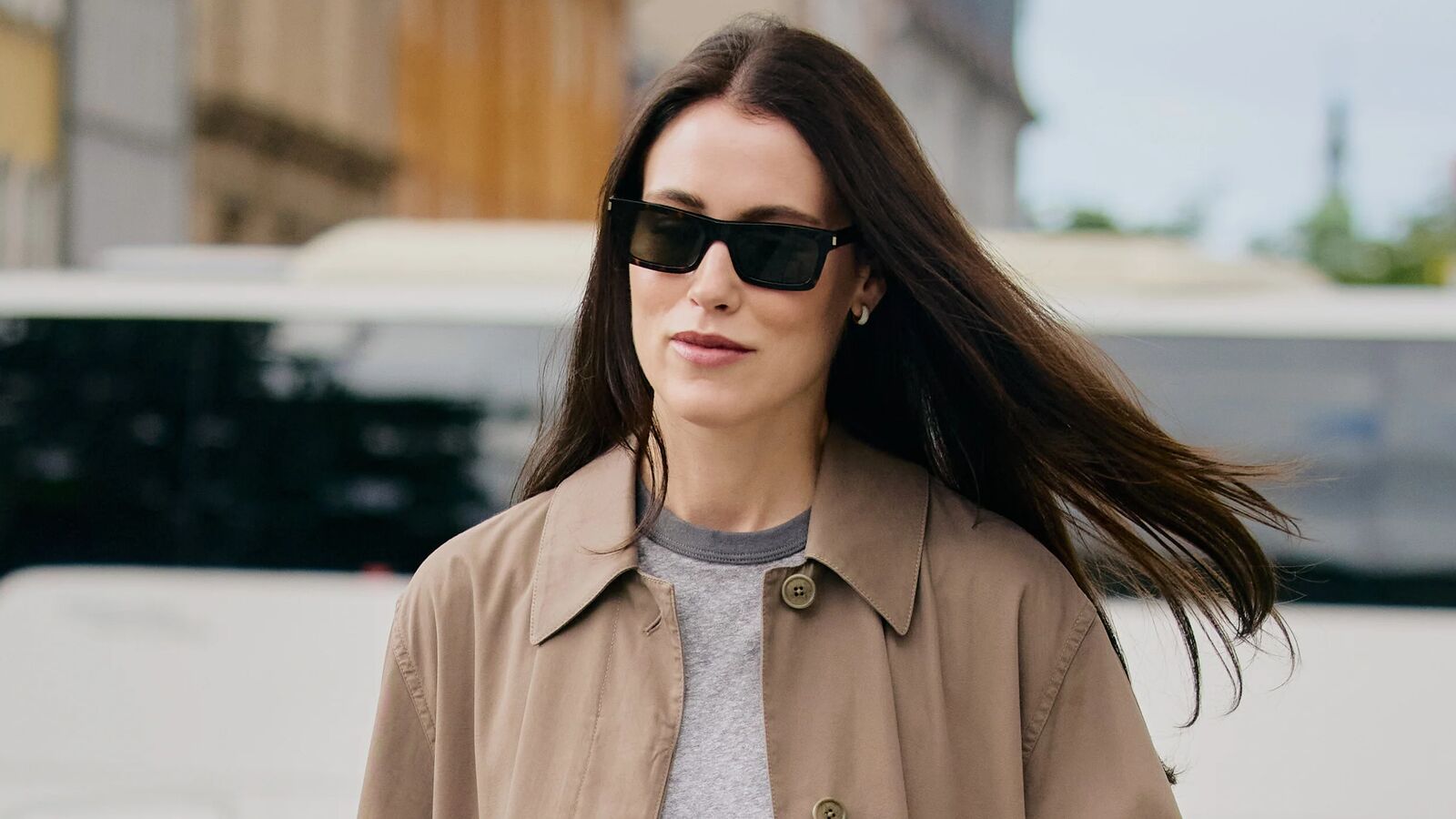 Woman wearing black rectangular sunglasses and a tan jacket over a gray crewneck top. 