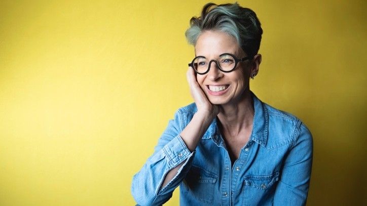 Portrait of Liz O&#039;Riordan, a white woman with short greying hair and round-framed glasses, against a yellow background. She is resting her face on one hand and smiling 