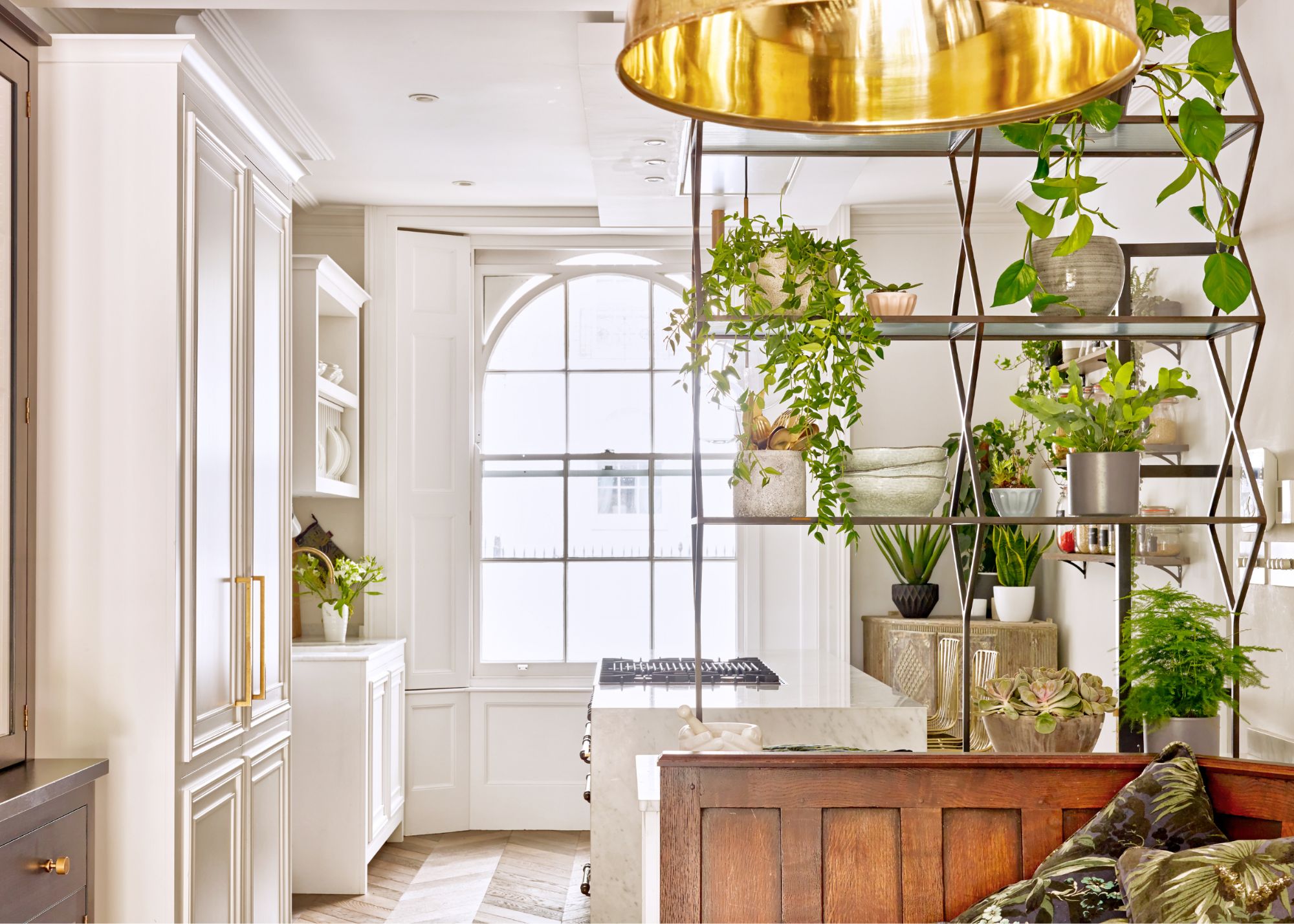 A kitchen dining area with a large gold pendant light hanging from above in front of a divider filled with small potted plants between the kitchen area with an island and some freestanding cabinetry