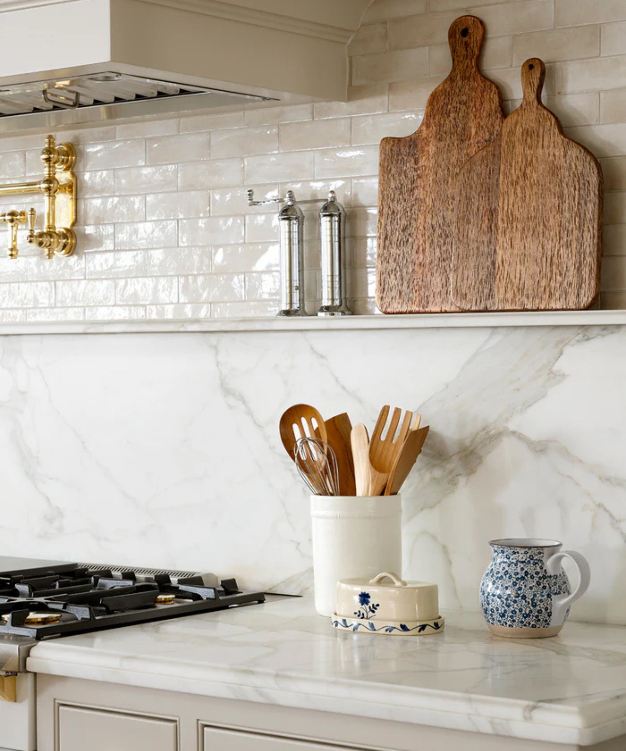 Marble countertop and backsplash with wooden chopping boards, utensil pot with wooden utensils, butter dish and blue and white floral jug