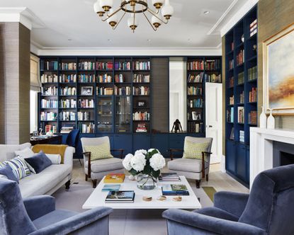Large family room space with floor to ceiling shelving painted blue, white ceiling and light wooden flooring, beige wallpapered walls, white fireplace, two gray lounge chairs with dark wood frames, gray sofa with black feet, two blue velvet lounge chairs, white square coffee table with books and flowers, white and metal lantern chandelier