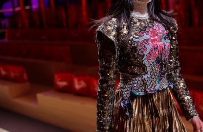 Model wearing highly-detailed coat with sequins and a dragon motif, with a pleated gold skirt