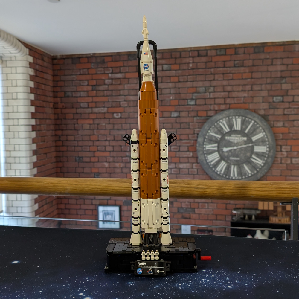An orange and white pointed rocket displayed against a brick wall with a large clock on it.