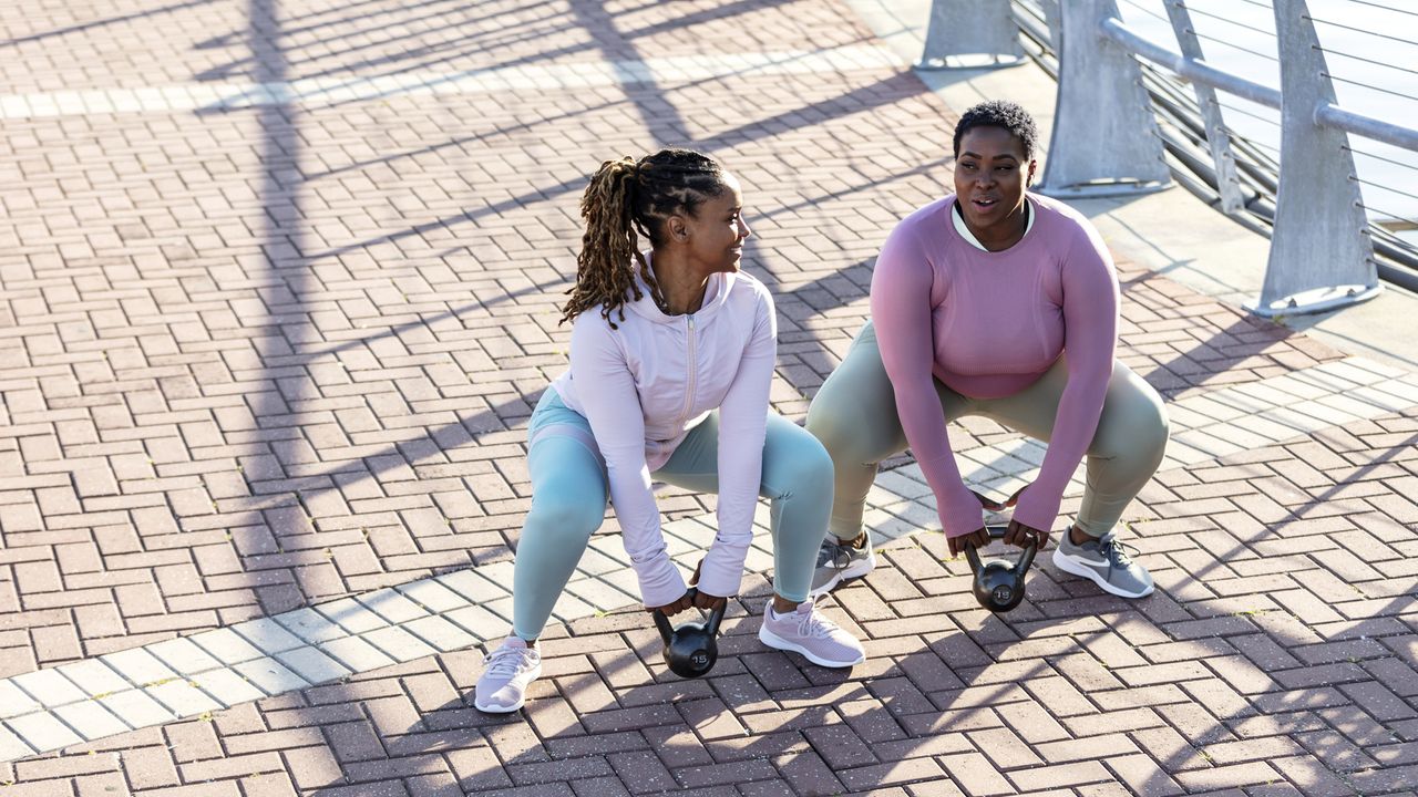 Two women exercising outside, squatting down to grab dumbbells on the ground in front of them