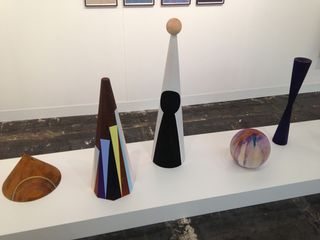 Five sculptures on display. Left to right: A shell style ornament. A cone style ornament with purple and green triangles painting on it. A cone with a ball at its top point. An apple style shape. An hour glass style shape.