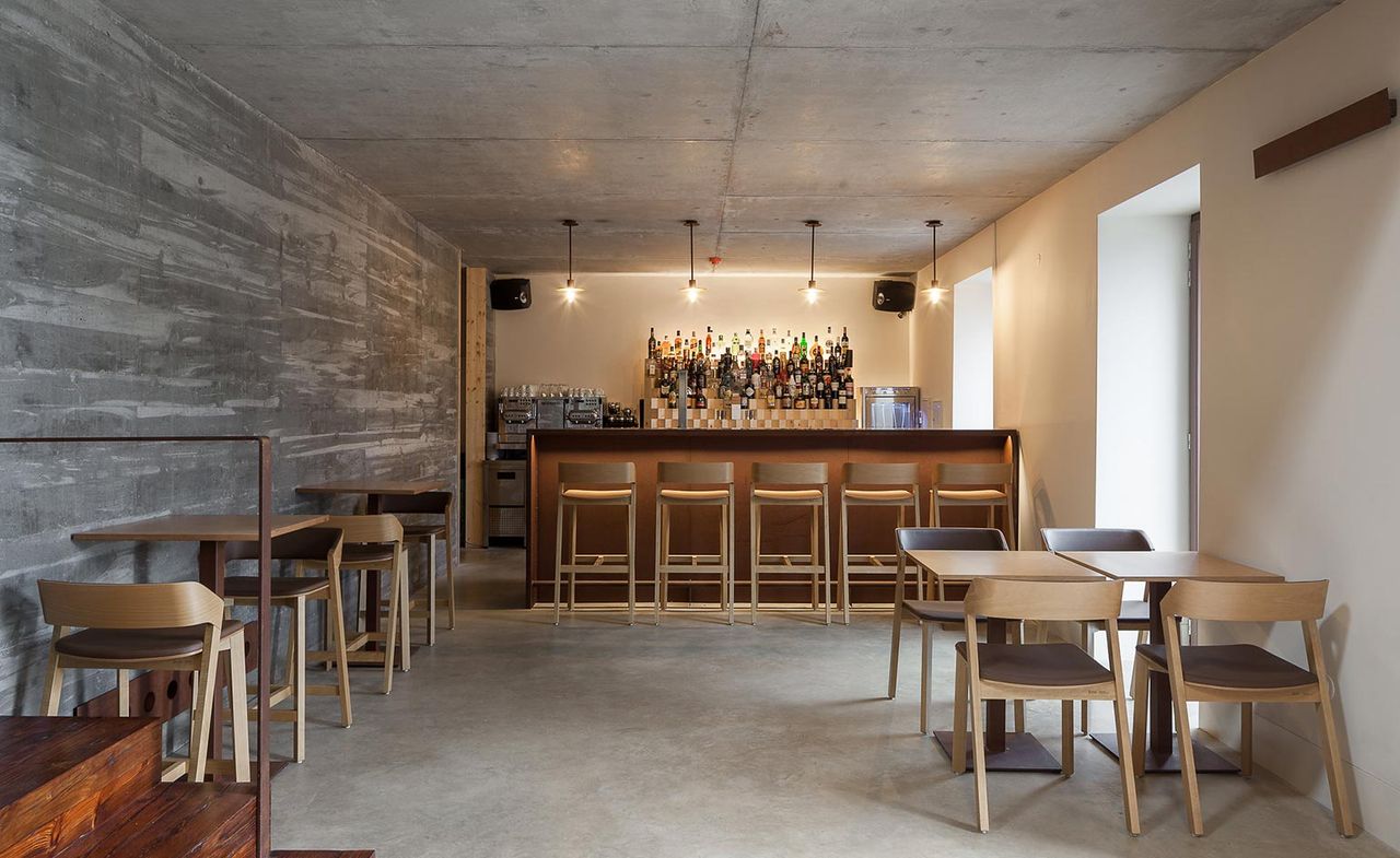 An interior view of the Entre Portas restaurants. Floors, walls, and ceilings are all in gray concrete syle. The bar brown color is on the far wall, and we see bottles of drinks behind it. The tables and chairs are all in light wood.