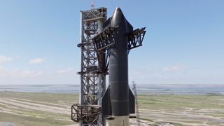 closeup of a black, cylindrical spacecraft next to its gray launch tower, with greenish-brown wetlands and the sea in the background.