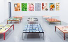 A series of bold and colourful tables in a white room which has bold prints on the walls
