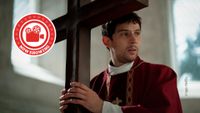Josh O'Connor holds a large crucifix in his hands while dressed in a deep red priest's robe. He looks off to the right of the image with an expression of quiet concern.