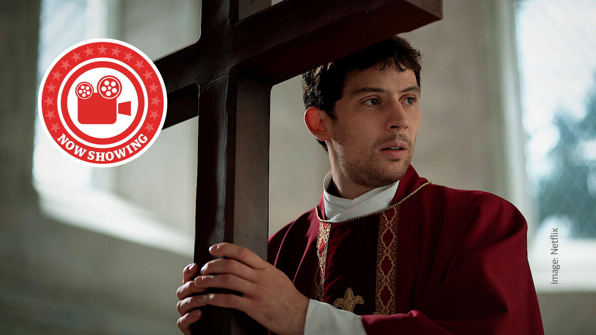 Josh O'Connor holds a large crucifix in his hands while dressed in a deep red priest's robe. He looks off to the right of the image with an expression of quiet concern.