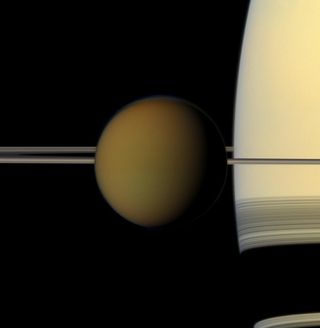 A brown/yellow sphere of Titan is in the center of the image. Sandy yellow Saturn is behind.