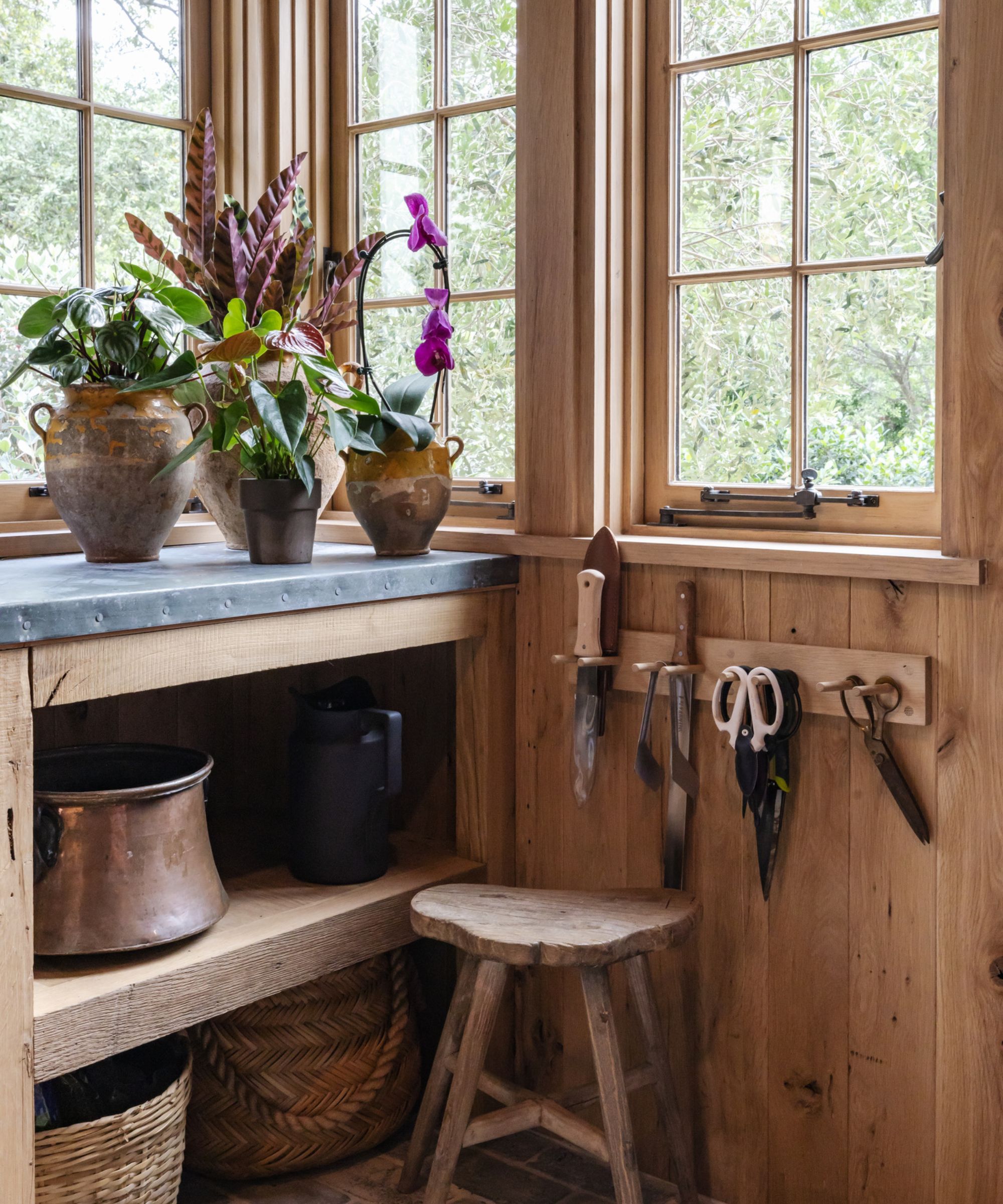 The corner of a shed with large windows. A potting bench with plants on top, a small stool beside it, and a small wall rack with shears hanging from it.