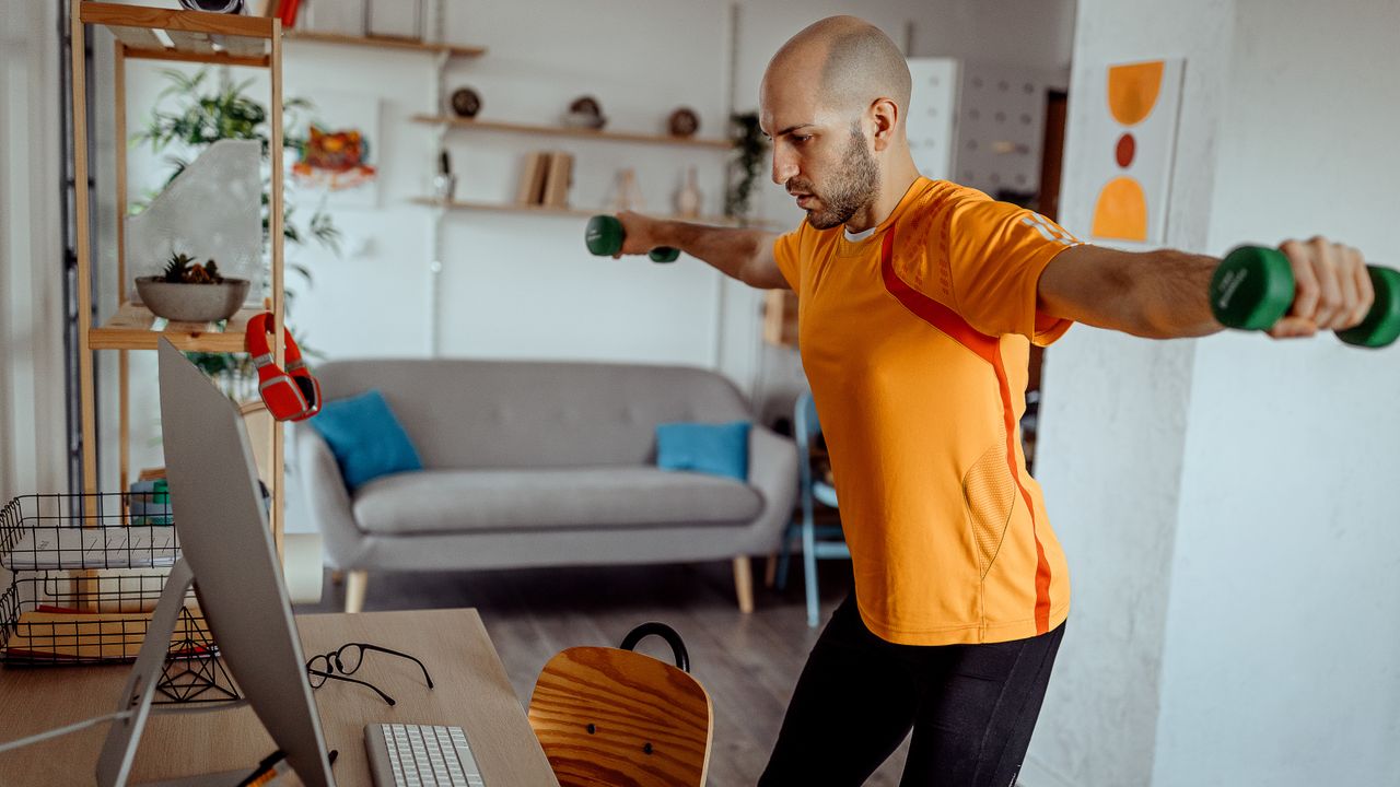 A man stands in an orange t-shirt and leggings and performs a dumbbell lateral raise in a small apartment. Behind him we can see a sofa, some shelves and plants. Directly in front of him is a computer screen and keyboard. He holds light dumbbells in his hands and raises them out the sides, keeping his arms straight.