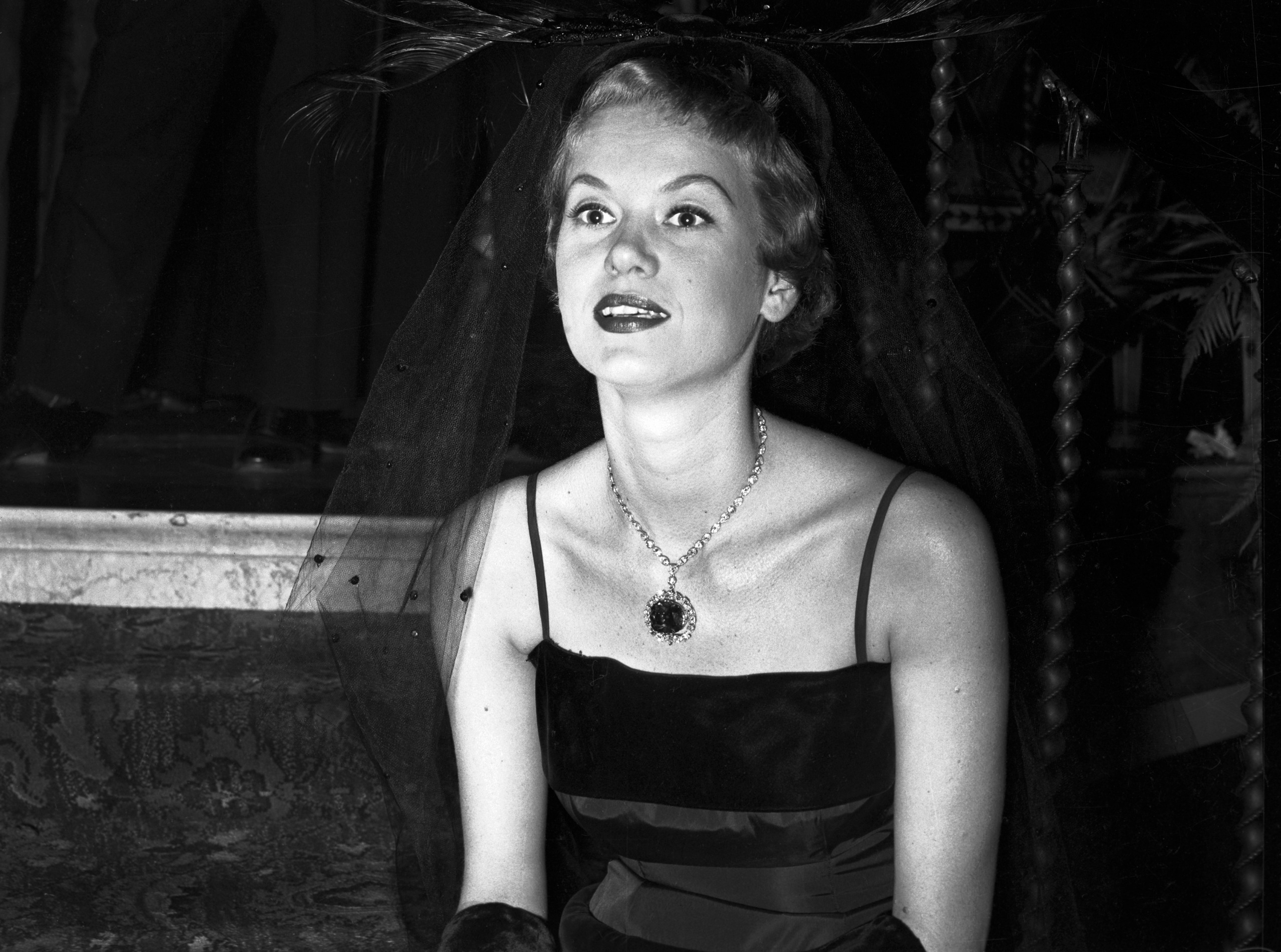 A black-and-white photograph of a woman in a square-neck, strapless velvet dress. She's wearing a very large, blue diamond necklace and a black netted headpiece