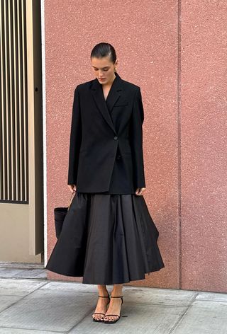 a photo showing the fall jacket trend, hourglass blazer worn by a woman standing against a pink wall wearing a black nipped waist blazer with a black skirt and black strappy sandals