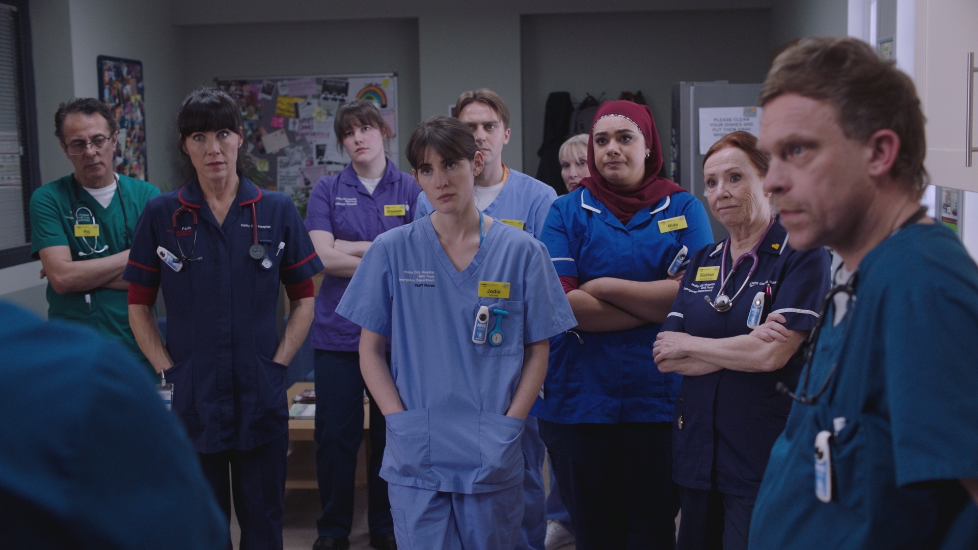 A group shot of the Casualty medics being gathered for a serious announcement in the staff room.