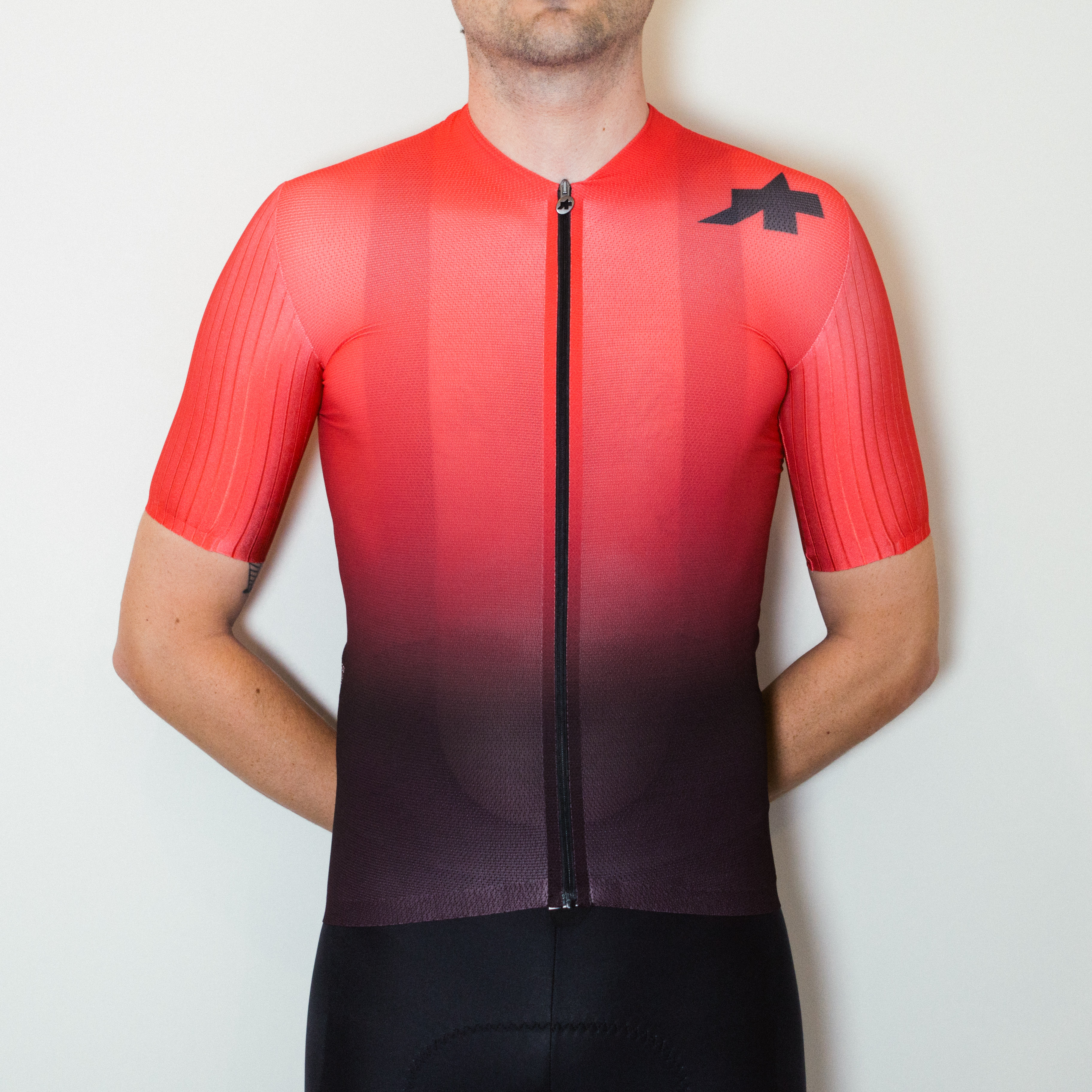 A white man in a red to black fade cycling jersey and black cycling shorts stands against a white wall