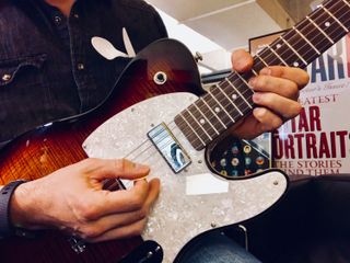 A human holds a guitar while sitting in an office cubicle on the East Coast of the United States. He has plastic cutlery in his shirt pocket. No one knows why.