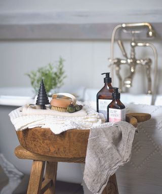 Wooden bowl with bottles, towels, brush on a wooden stool with a grey roll top bath in the background.