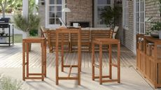 A patio with IKEA's NAMARRO bar table and two bar stools with a lamp, mug and laptop on top of the table, next to an outdoor console, with a seating area in the background and a bbq next to the grass