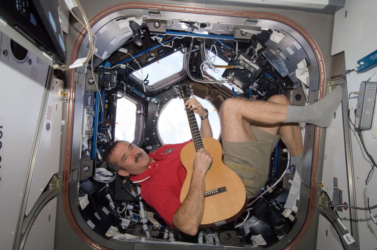 chris hadfield holding a guitar and backdropped by a wraparound window in space