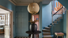 entryway with mid-blue walls, black and white floor tiles, round black table in centre with vase of flowers, stairway with bold artwork on the wall