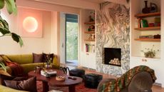 A living room with an olive green couch, a pair of black poufs, a brown sofa seater, a wood coffee table, a marble fireplace, and floating shelves with books, ceramics, and decor